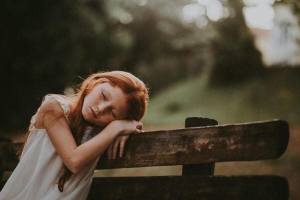 A young girl sleeps peacefully on a wooden bench outdoors, surrounded by nature.