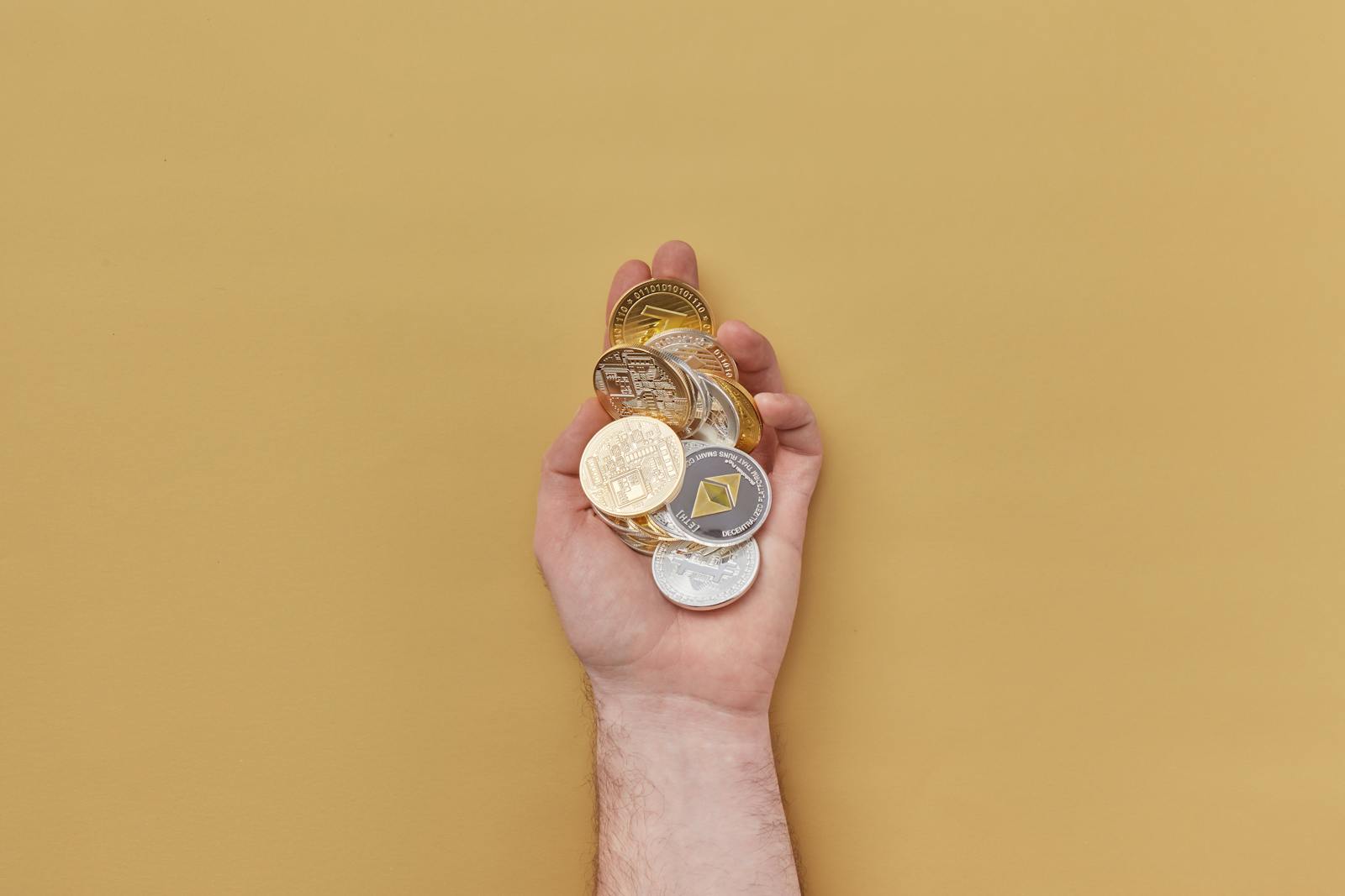 Close up of a hand holding various cryptocurrency coins against a yellow background